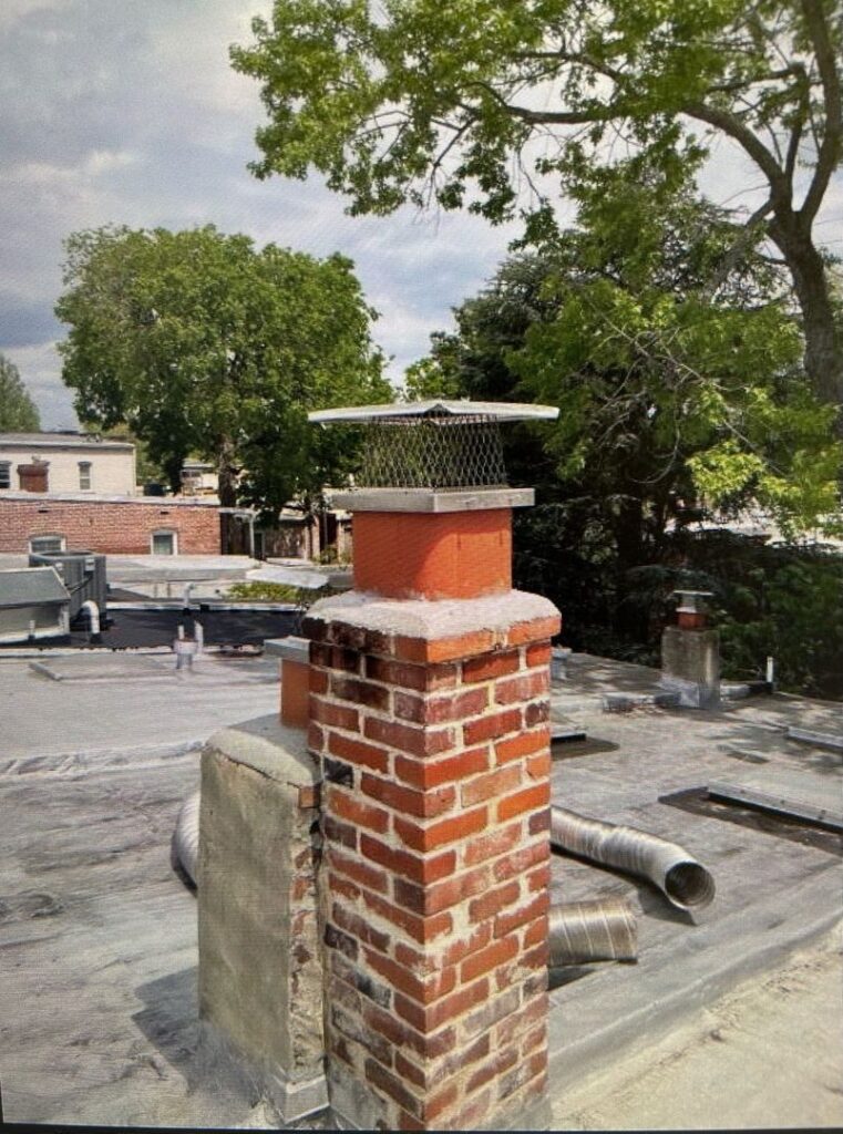 Brick chimney with a metal cap on a flat rooftop, metal ducts scattered nearby, and an urban landscape in the background.