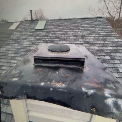 A photo showing a damaged and rusty chimney chase cover on a house roof with shingles and another chimney visible in the background