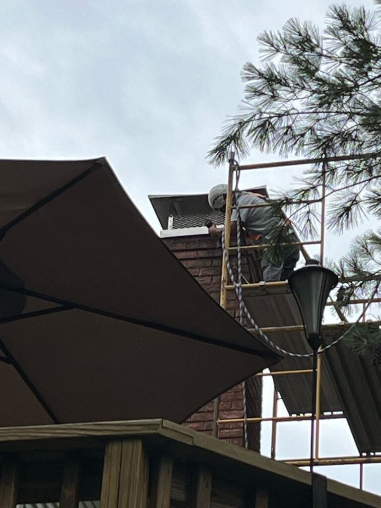 A worker installs a mount cap on a brick chimney using scaffolding in an outdoor residential area.