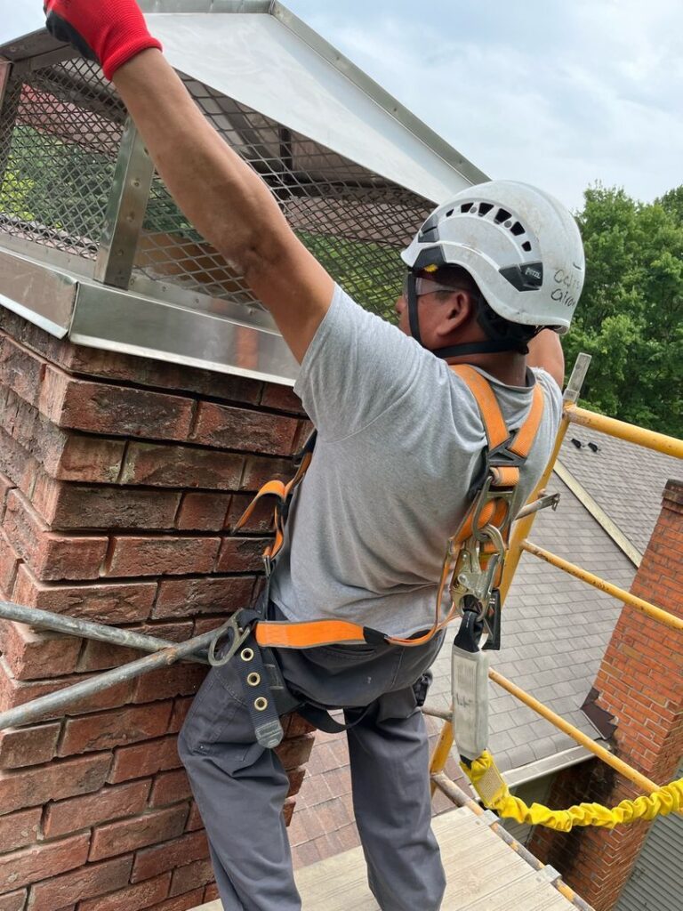 A worker wearing safety gear installs a metal cap on a brick chimney while standing on scaffolding.